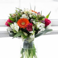 An exquisite arrangement, reminiscent of the floral artistry found along Romford's bustling South Street, sits proudly in a gleaming glass vase. Cream and peach gerbera daisies form a soft halo at the center, their delicate petals fanning outward to reveal glowing heart-like discs in brilliant orange and chestnut brown. Vibrant fuchsia roses, their spiral centers lush and inviting, provide a sumptuous depth that hints at romance and thoughtful craftsmanship-attributes cherished by the Flowers Romford team. Intermingled with these focal flowers are sprays of snowy white alstroemeria and petite white rosebuds, lending gentle brightness and texture that recall quiet strolls through Lodge Farm Park. The ensemble is anchored by swathes of richly coloured aspidistra leaves; these gracefully wrap the bouquet base, both supporting the structure and infusing the piece with earthy tranquility. Narrow, pointed greens and fairy-like wisps of foliage dart amongst the blooms, amplifying volume and visual interest. Through the vase's crystal walls, the orderly green stems are seen standing tall, symbolizing the freshness and vitality that define professional floristry here in Romford. Illuminated against a soft, neutral backdrop, this bouquet radiates joy and connection-its optimistic colours and harmonious structure making it the perfect centrepiece for shared celebrations, heartfelt congratulations, or a quiet moment of gratitude.